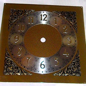 May include: A square clock face plate featuring a central circular dial with metallic numerals from 1 to 12. The background is a textured brown finish, accented by ornate, raised gold-toned corner embellishments and a circular center hole.