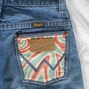 May include: Blue denim jeans with a back pocket featuring a colorful abstract design and a brown leather patch with the word "Wrangler" printed on it. The jeans also have a brown leather patch with the word "Wrangler" printed on it above the back pocket.