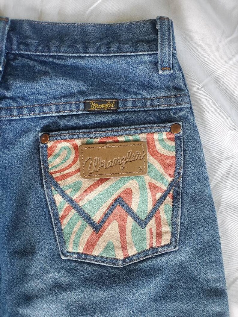May include: Blue denim jeans with a back pocket featuring a colorful abstract design and a brown leather patch with the word "Wrangler" printed on it. The jeans also have a brown leather patch with the word "Wrangler" printed on it above the back pocket.