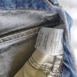 May include: A close-up of a blue denim jean label with the text "13MWZBP 16 REG 16 COUR. LAS-D282-02 510713" printed on it. The label is made of white fabric and has a white background.