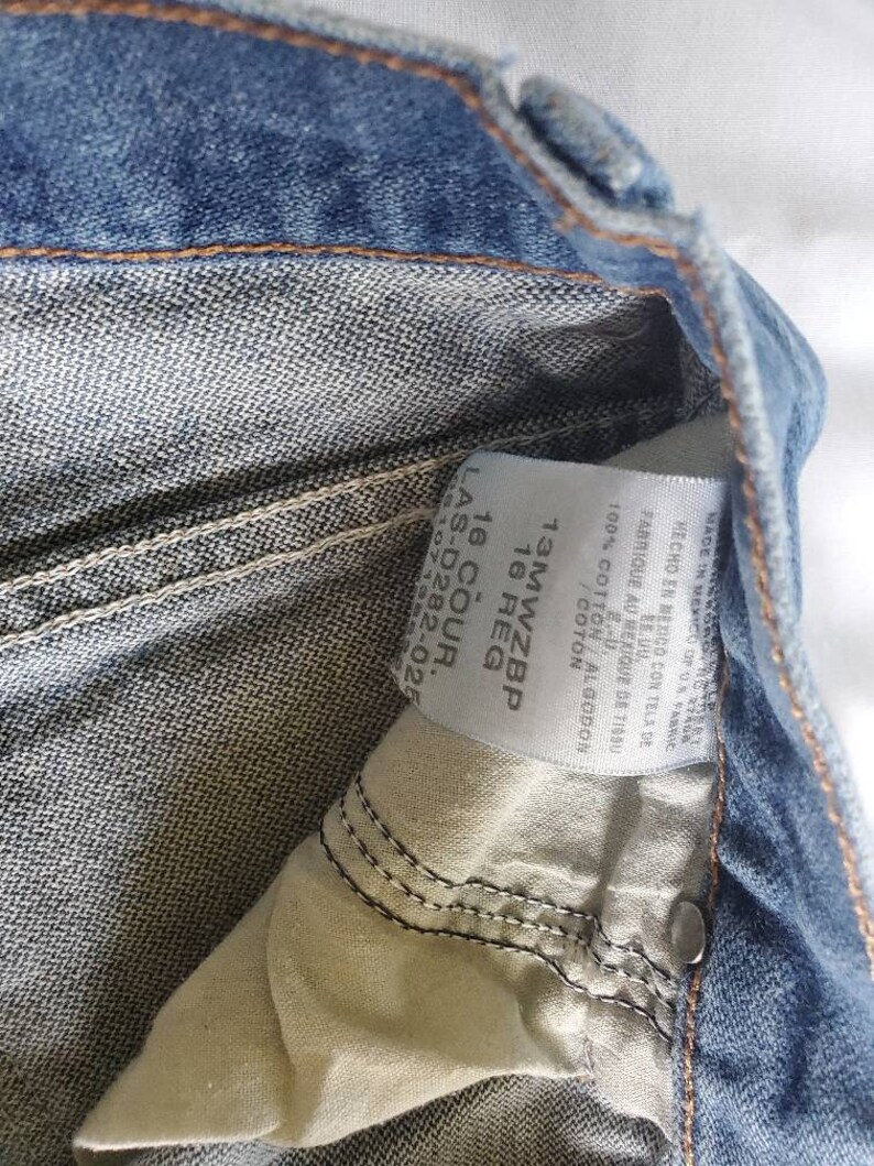 May include: A close-up of a blue denim jean label with the text "13MWZBP 16 REG 16 COUR. LAS-D282-02 510713" printed on it. The label is made of white fabric and has a white background.