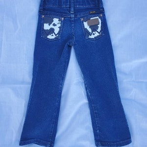 May include: Blue denim jeans with a bootcut leg opening. The back pockets have a cowhide print with a brown leather patch that says "Wrangler" in white.