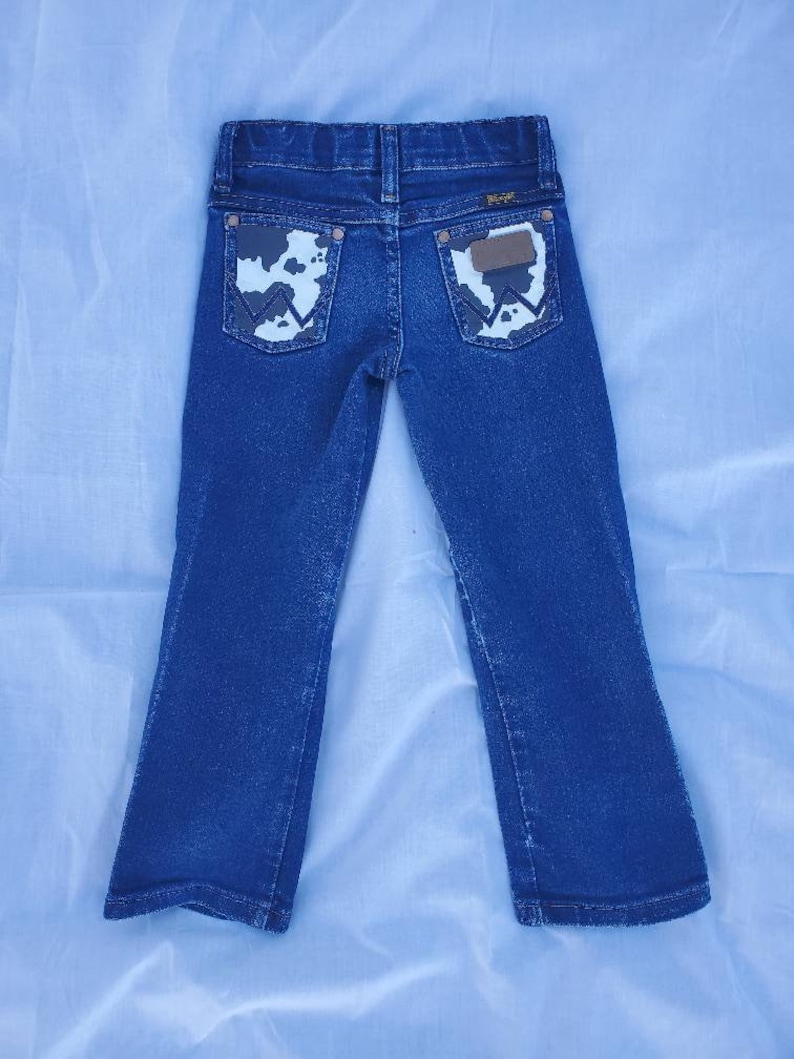May include: Blue denim jeans with a bootcut leg opening. The back pockets have a cowhide print with a brown leather patch that says "Wrangler" in white.