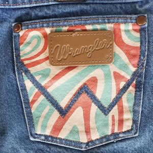 May include: A pair of blue denim jeans with a back pocket featuring a red, white, and green abstract swirl pattern. The pocket has a brown leather patch with the word "Wrangler" in cursive.