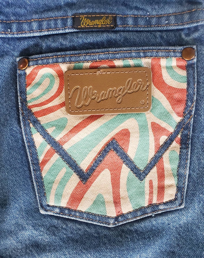 May include: A pair of blue denim jeans with a back pocket featuring a red, white, and green abstract swirl pattern. The pocket has a brown leather patch with the word "Wrangler" in cursive.