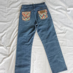 May include: A pair of blue denim jeans with a unique design on the back pockets. The pockets have a swirling pattern in shades of orange, green, and blue.