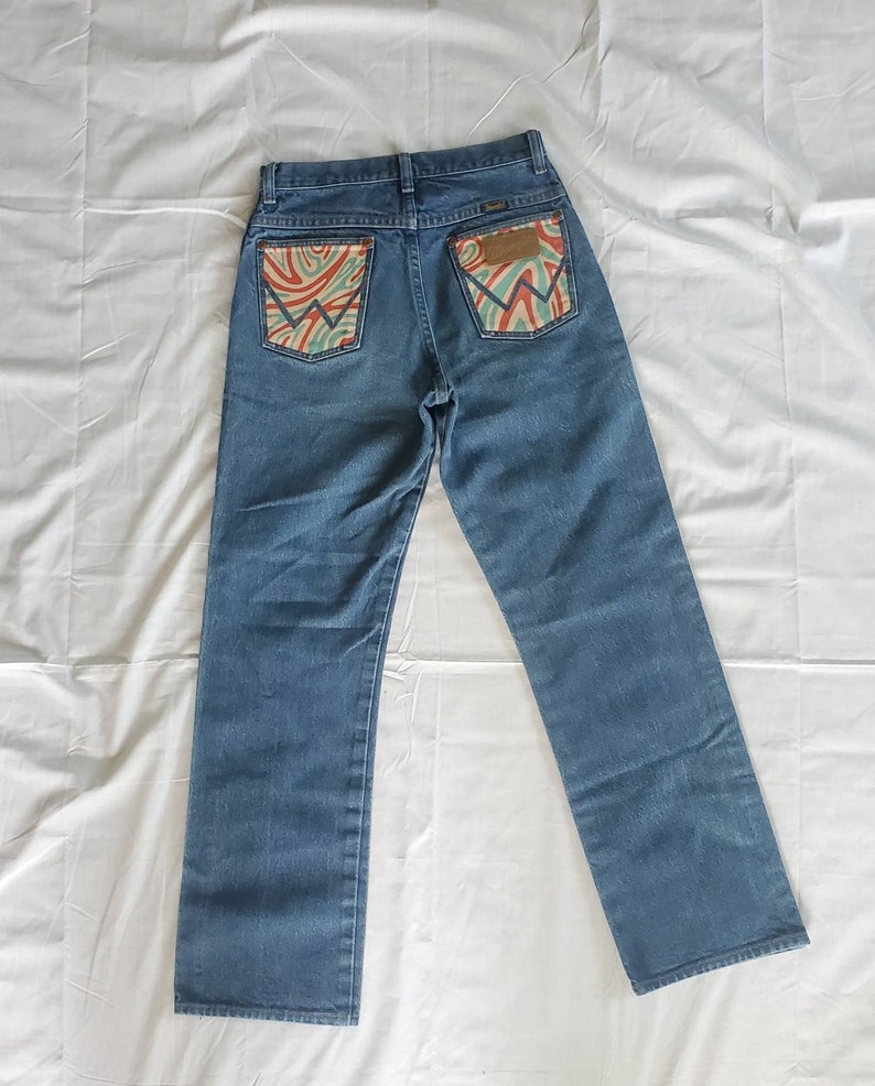 May include: A pair of blue denim jeans with a unique design on the back pockets. The pockets have a swirling pattern in shades of orange, green, and blue.