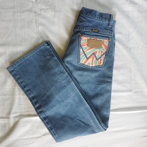 May include: A pair of blue denim jeans with a back pocket featuring a colorful, abstract design and the Wrangler logo.