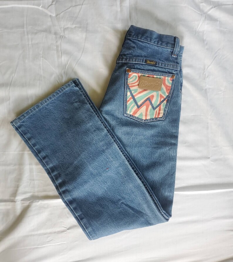 May include: A pair of blue denim jeans with a back pocket featuring a colorful, abstract design and the Wrangler logo.