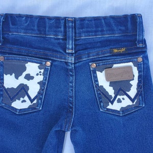 May include: A pair of blue denim jeans with a cow print patch on the back pockets. The patch has the Wrangler logo on it.