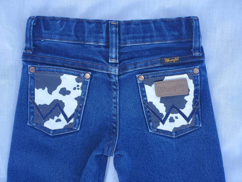 May include: A pair of blue denim jeans with a cow print patch on the back pockets. The patch has the Wrangler logo on it.