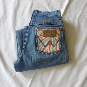 May include: A pair of blue denim jeans with a back pocket featuring a colorful, abstract design and a brown leather Wrangler label.