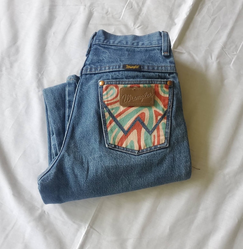 May include: A pair of blue denim jeans with a back pocket featuring a colorful, abstract design and a brown leather Wrangler label.