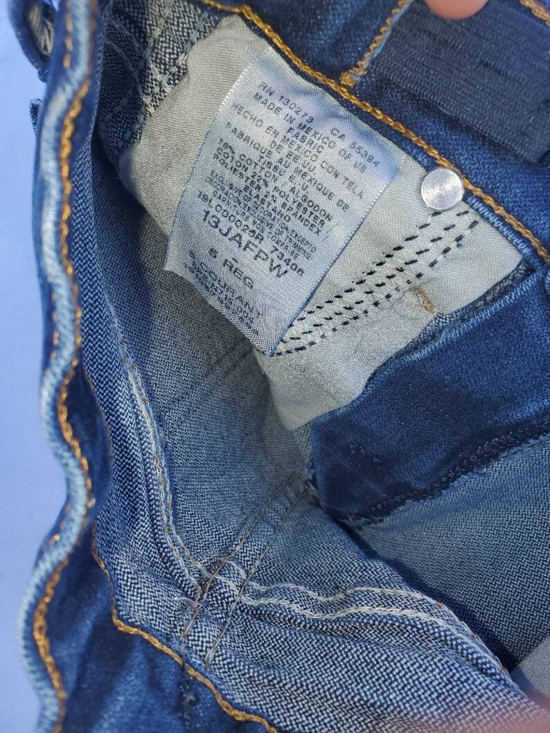 May include: A close-up of a blue denim jean label with white text. The label includes the text "RN 130273 CA 55384 MADE IN MEXICO OF US FABRIC HECHO EN MEXICO CON TELA DE RE.UU. FABRIQUE AU MEXIQUE DE TISSU EU. 78% COTTON / ALGODON 22% POLYESTER POLIESTER 2% BRANDEX ELASTANO 1910000268 73406 13JAFPW S REG".
