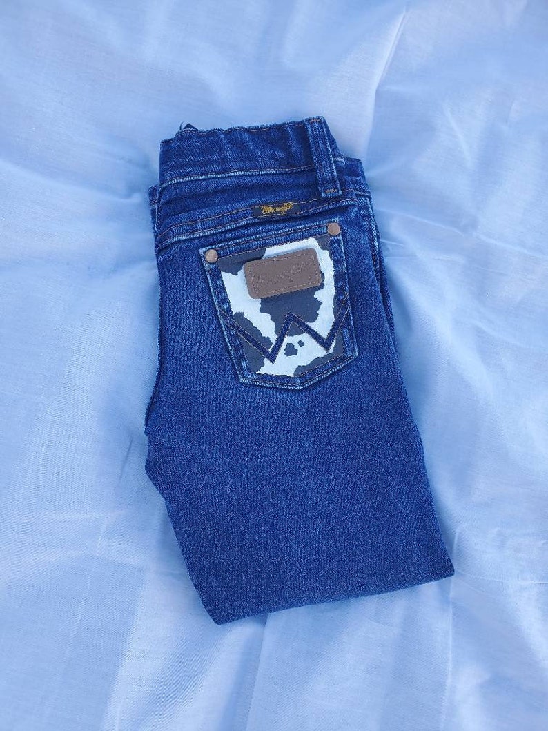 May include: A pair of blue denim jeans with a cowhide patch on the back pocket. The patch has a white and black pattern and the Wrangler logo is visible.