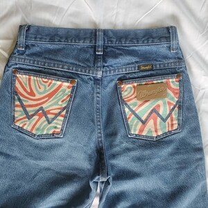 May include: A pair of blue denim jeans with a unique design on the back pockets. The pockets have a swirling pattern in shades of red, green, and white. The Wrangler logo is visible on the right pocket.