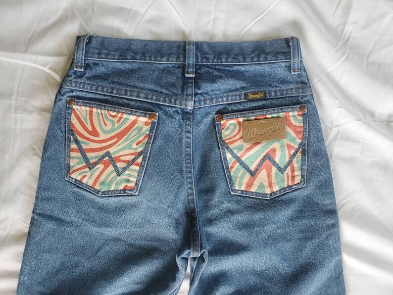 May include: A pair of blue denim jeans with a unique design on the back pockets. The pockets have a swirling pattern in shades of red, green, and white. The Wrangler logo is visible on the right pocket.