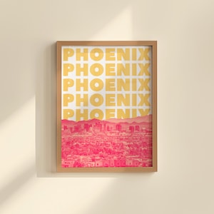 May include: A framed art print featuring the city of Phoenix. The print has the word "PHOENIX" in a repeating pattern in yellow over a pink and red cityscape. The frame is made of light brown wood.