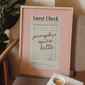 May include: A framed print of a vintage guest check with the words "pumpkin spice latte" in cursive script. The background has a pink and white striped design. A cup of coffee on a saucer is in the foreground.
