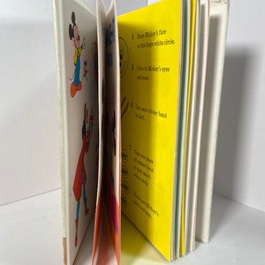 May include: A children's book with instructions on how to make a Mickey Mouse puppet. The book has colourful illustrations of Mickey Mouse and instructions on how to draw his face, colour his eyes and nose, cut rubber bands in half, and tape them to small white circles.