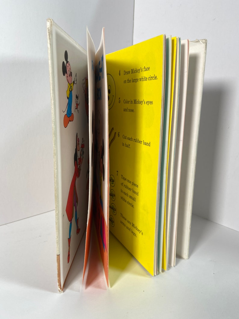 May include: A children's book with instructions on how to make a Mickey Mouse puppet. The book has colourful illustrations of Mickey Mouse and instructions on how to draw his face, colour his eyes and nose, cut rubber bands in half, and tape them to small white circles.