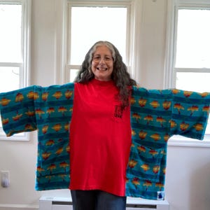 Pu&ograve; includere: Una persona indossa un kimono turchese con un motivo di forme gialle e arancioni. &Egrave; indossato sopra una maglietta rossa e jeans blu. Il kimono ha delle tasche.