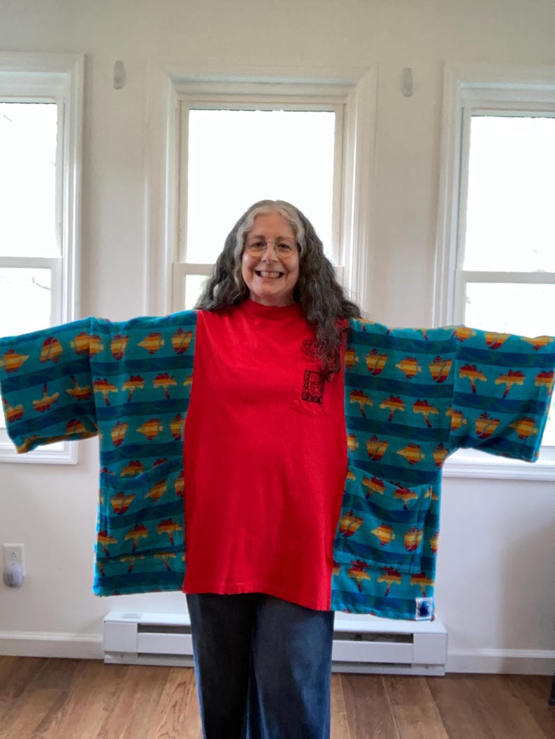 Pu&ograve; includere: Una persona indossa un kimono turchese con un motivo di forme gialle e arancioni. &Egrave; indossato sopra una maglietta rossa e jeans blu. Il kimono ha delle tasche.