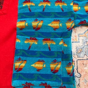 Pu&ograve; includere: Una maglietta rossa a maniche corte con una tasca con testo giapponese e un accappatoio turchese con un motivo tropicale. L'accappatoio presenta un motivo ripetuto di palme e pesci.