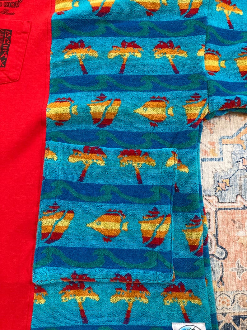 Pu&ograve; includere: Una maglietta rossa a maniche corte con una tasca con testo giapponese e un accappatoio turchese con un motivo tropicale. L'accappatoio presenta un motivo ripetuto di palme e pesci.