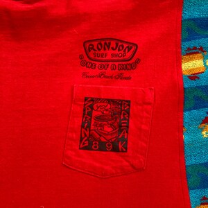 Pu&ograve; includere: Una maglietta rossa senza maniche con una tasca con una grafica nera. La grafica include il testo "SPRING BREAK 89K" e un disegno di una persona. La maglietta ha anche il testo "RONJON SURF SHOP ONE OF A KIND Cocoa Beach Florida".
