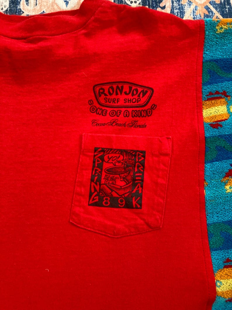 Pu&ograve; includere: Una maglietta rossa senza maniche con una tasca con una grafica nera. La grafica include il testo "SPRING BREAK 89K" e un disegno di una persona. La maglietta ha anche il testo "RONJON SURF SHOP ONE OF A KIND Cocoa Beach Florida".