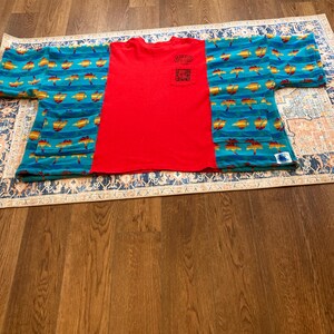 Pu&ograve; includere: Una maglietta rossa con taschino &egrave; abbinata a un top stile kimono blu e giallo fantasia. Il kimono presenta un motivo ripetuto di forme gialle e arancioni su sfondo blu. La maglietta e il kimono sono esposti su un tappeto fantasia.