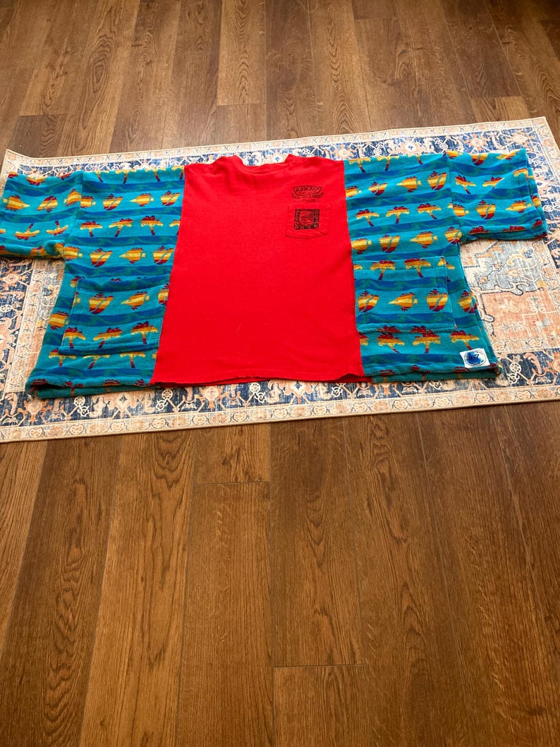 Pu&ograve; includere: Una maglietta rossa con taschino &egrave; abbinata a un top stile kimono blu e giallo fantasia. Il kimono presenta un motivo ripetuto di forme gialle e arancioni su sfondo blu. La maglietta e il kimono sono esposti su un tappeto fantasia.