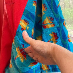 Pu&ograve; includere: Una camicia rossa con tessuto blu a fantasia tipo asciugamano sulle maniche e sulla tasca. Il tessuto dell'asciugamano presenta forme gialle, arancioni e rosse su sfondo blu. Una mano tiene aperta la tasca.