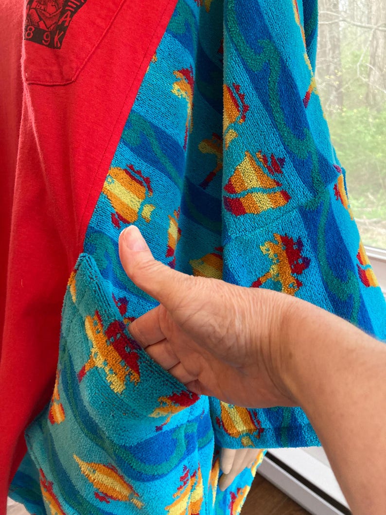 Pu&ograve; includere: Una camicia rossa con tessuto blu a fantasia tipo asciugamano sulle maniche e sulla tasca. Il tessuto dell'asciugamano presenta forme gialle, arancioni e rosse su sfondo blu. Una mano tiene aperta la tasca.