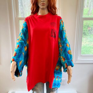 Pu&ograve; includere: Una maglietta rossa a maniche lunghe con una tasca e un logo. Le maniche sono decorate con forme blu, gialle e arancioni. La maglietta &egrave; abbinata a jeans blu strappati.