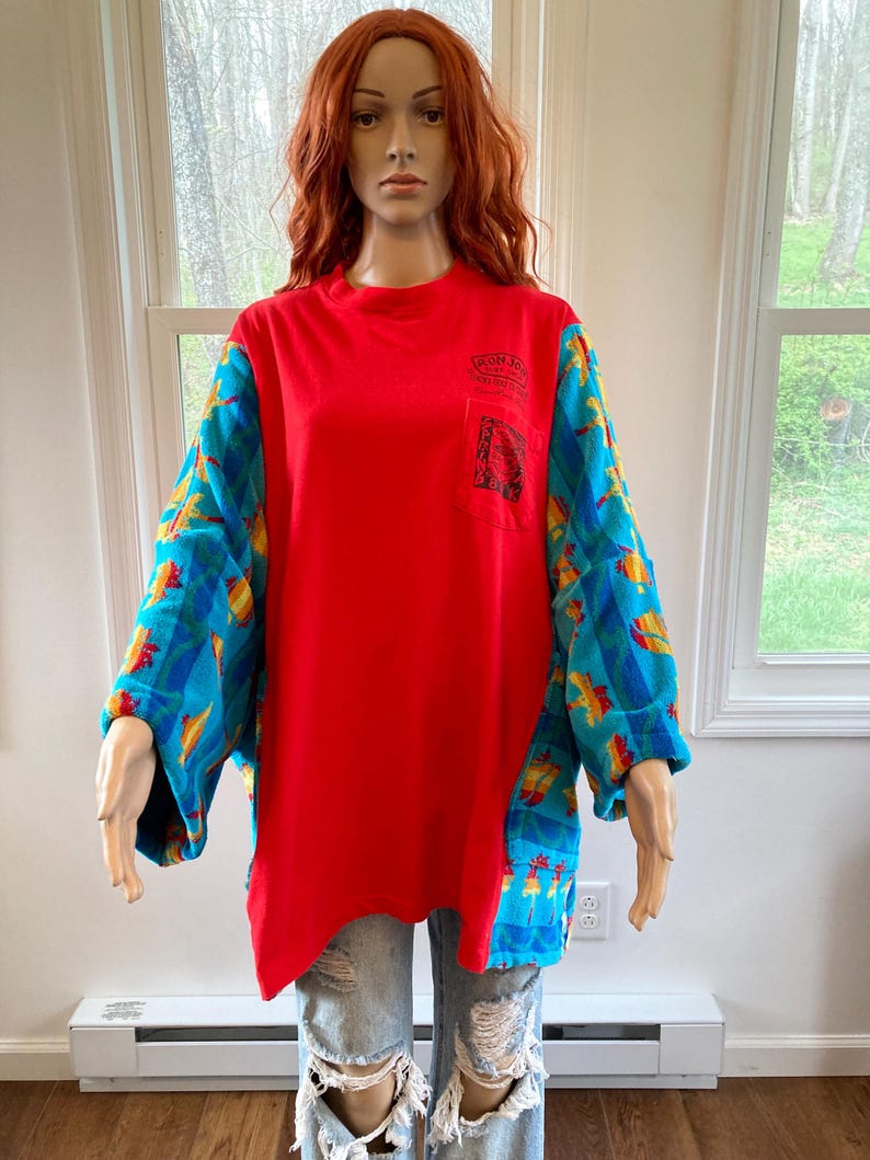 Pu&ograve; includere: Una maglietta rossa a maniche lunghe con una tasca e un logo. Le maniche sono decorate con forme blu, gialle e arancioni. La maglietta &egrave; abbinata a jeans blu strappati.