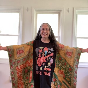 Puede incluir: Una camiseta negra con el texto "The Monkees GOOD TIME" se usa debajo de un chal acolchado estampado. El chal presenta una mezcla de colores rojo, verde y amarillo con diseños de cachemira y geométricos. El fondo muestra tres ventanas.