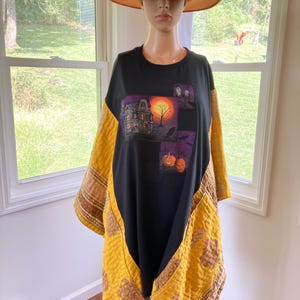 Puede incluir: Una camiseta negra con un estampado gráfico de una casa encantada, una luna llena y calabazas, acentuada con paneles de tela acolchada de color amarillo mostaza en los laterales y mangas, exhibida en un maniquí con un sombrero naranja.