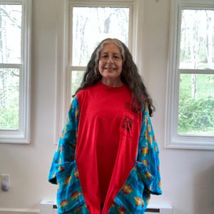 Pu&ograve; includere: Una camicia patchwork rossa e blu con maniche lunghe e grandi tasche. La camicia ha un pannello centrale rosso e maniche e tasche blu fantasia. Il motivo include forme gialle e arancioni. La camicia &egrave; indossata da una persona.