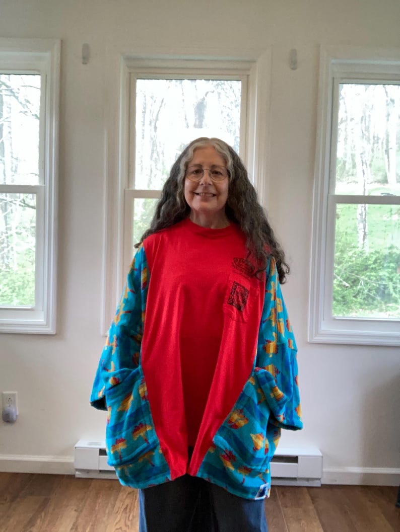 Pu&ograve; includere: Una camicia patchwork rossa e blu con maniche lunghe e grandi tasche. La camicia ha un pannello centrale rosso e maniche e tasche blu fantasia. Il motivo include forme gialle e arancioni. La camicia &egrave; indossata da una persona.