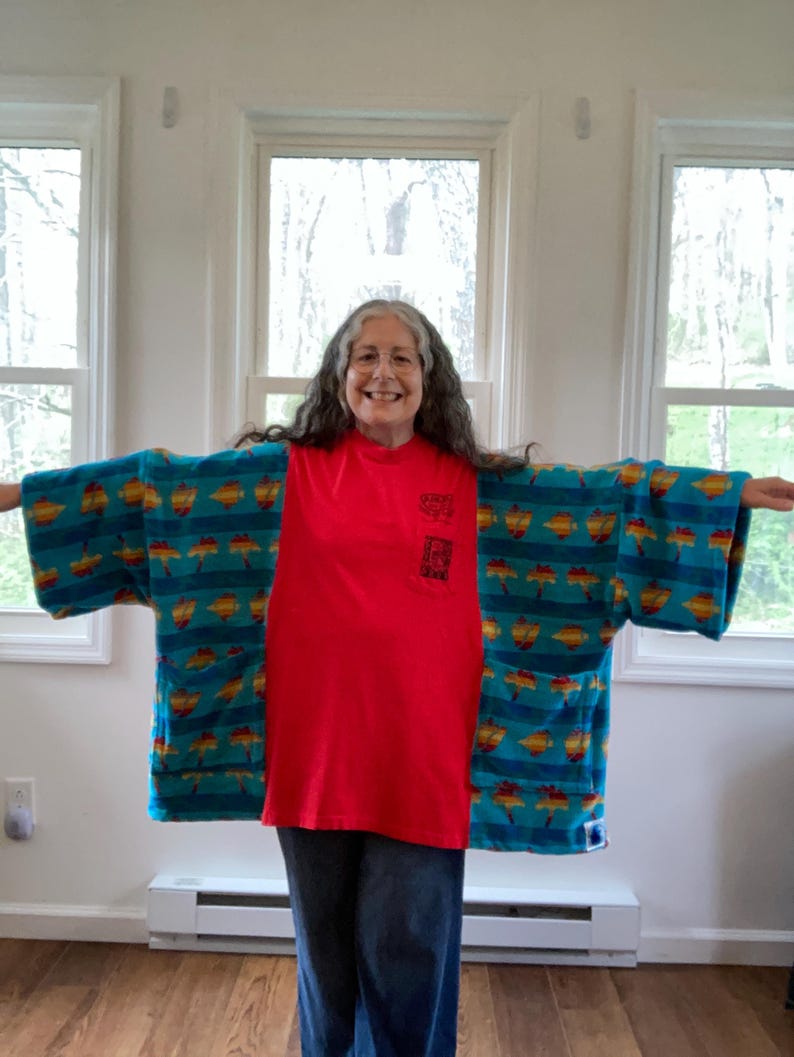 Pu&ograve; includere: Una maglietta rossa con una piccola grafica sulla tasca &egrave; indossata sotto un kimono a fantasia turchese e giallo. Il kimono presenta un motivo ripetuto di forme gialle e arancioni su sfondo turchese. La persona indossa jeans blu.