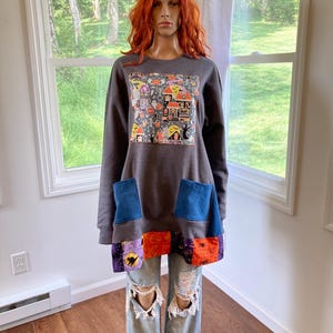 Puede incluir: Una sudadera de manga larga en color gris carbón con un parche cuadrado central que presenta un estampado colorido de temática de Halloween con casas, fantasmas y calabazas. Incluye dos bolsillos de mezclilla azul y un dobladillo de patchwork.