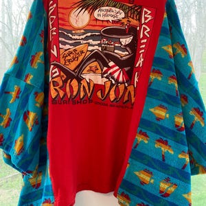 Pu&ograve; includere: T-shirt rosso con una grafica di una scena sulla spiaggia, palme e la scritta "Another Day in Paradise". Le maniche sono realizzate con un asciugamano blu fantasia con disegni di pesci e palme. La maglietta ha anche la scritta "Ron Jon Surf Shop Cocoa Beach, FLA."