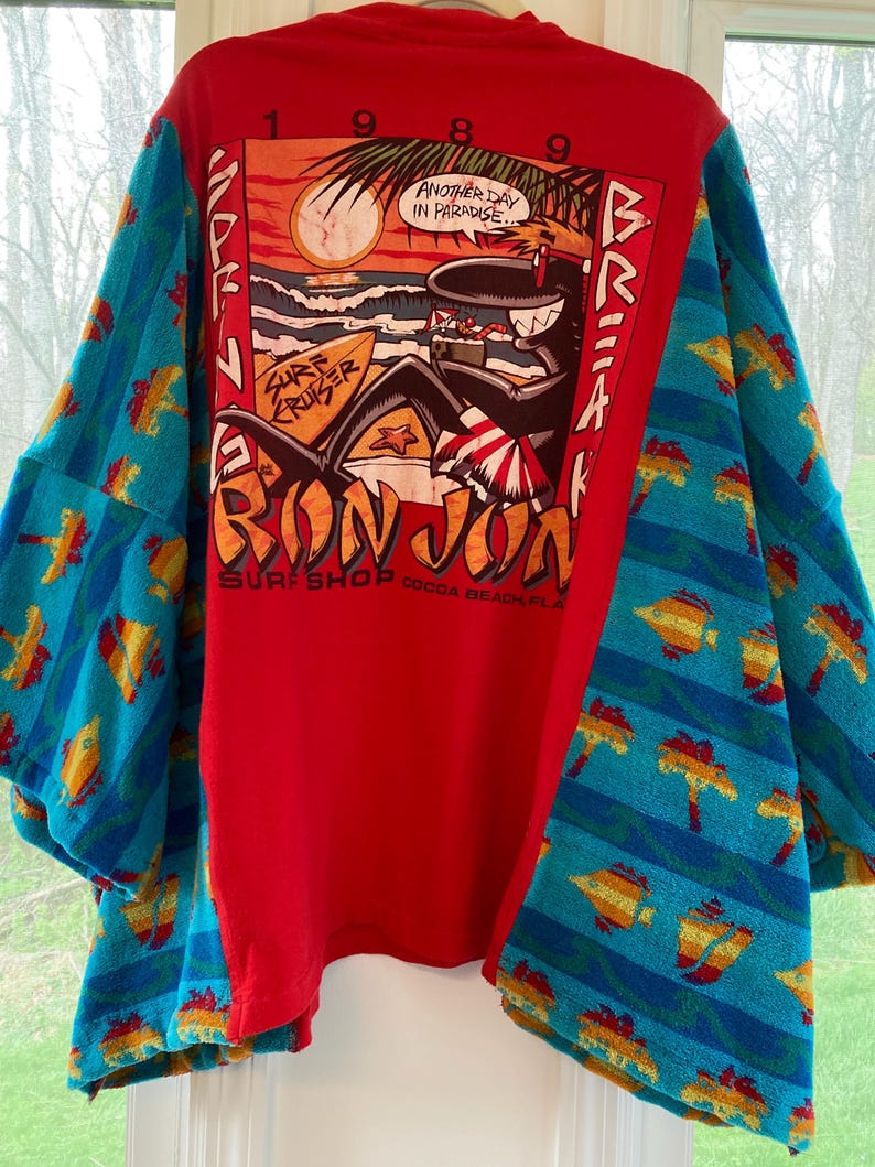 Pu&ograve; includere: T-shirt rosso con una grafica di una scena sulla spiaggia, palme e la scritta "Another Day in Paradise". Le maniche sono realizzate con un asciugamano blu fantasia con disegni di pesci e palme. La maglietta ha anche la scritta "Ron Jon Surf Shop Cocoa Beach, FLA."