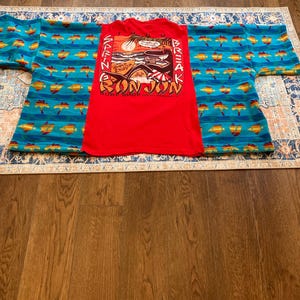 Pu&ograve; includere: Una maglietta rossa Bon Jon Surf Shop con una grafica di un tramonto, onde e una tavola da surf. La maglietta &egrave; appoggiata sopra un capo a fantasia blu e giallo. Lo sfondo &egrave; un pavimento in legno.