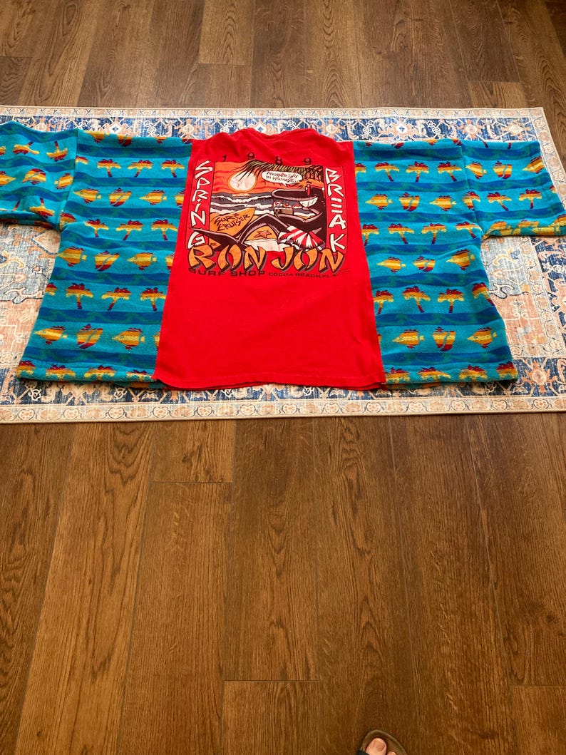 Pu&ograve; includere: Una maglietta rossa Bon Jon Surf Shop con una grafica di un tramonto, onde e una tavola da surf. La maglietta &egrave; appoggiata sopra un capo a fantasia blu e giallo. Lo sfondo &egrave; un pavimento in legno.