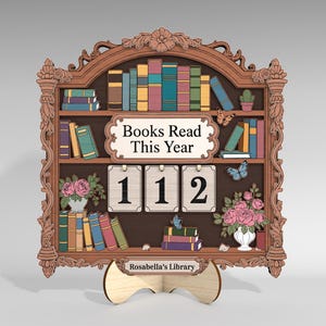 Könnte beinhalten: Ein holzgeschnitztes Bücherregal-Display mit dem Text "Books Read This Year" und der Zahl "112". Das Display zeigt Miniaturbücher, Blumen und Schmetterlinge. Der Boden trägt die Aufschrift "Rosabella's Library".