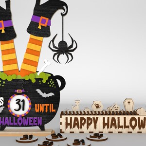 May include: A wooden Halloween countdown calendar with a witch's legs sticking out of a cauldron. The cauldron has a chalkboard with the text "Days Until Halloween" and a number wheel showing "31". There is a spider hanging from the cauldron. A separate wooden sign reads "Happy Halloween".