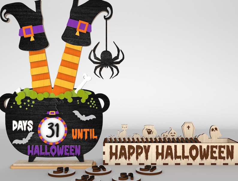 May include: A wooden Halloween countdown calendar with a witch's legs sticking out of a cauldron. The cauldron has a chalkboard with the text "Days Until Halloween" and a number wheel showing "31". There is a spider hanging from the cauldron. A separate wooden sign reads "Happy Halloween".